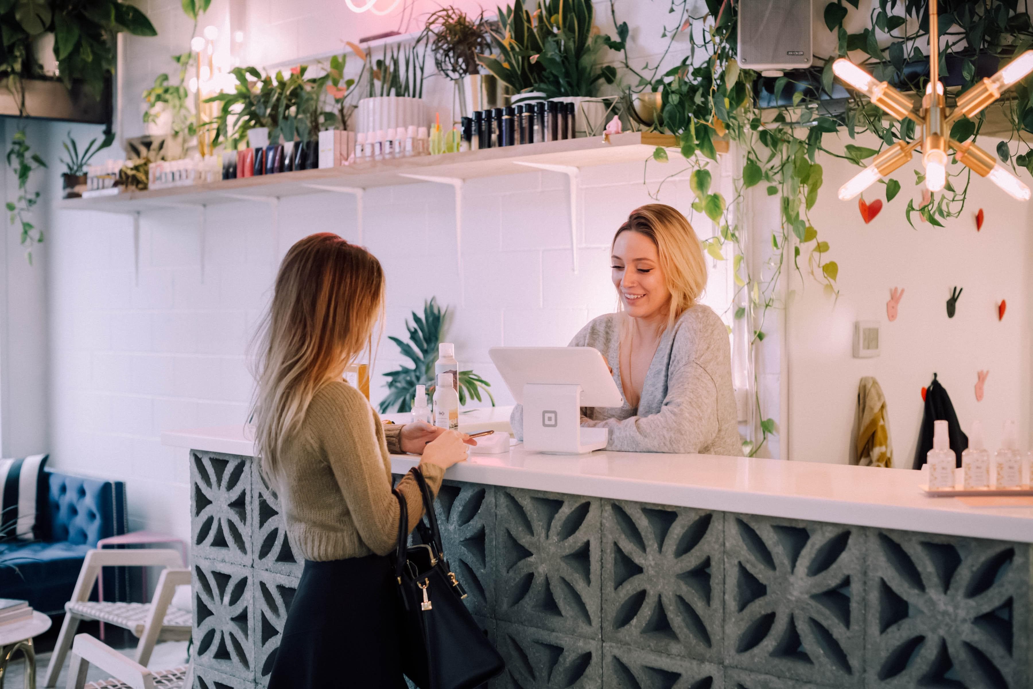A front-desk employee helps a customer at the counter of a plant-filled shop. NovaDesq status cards float over the scene: an incoming call answered, an appointment booked, the owner notified.